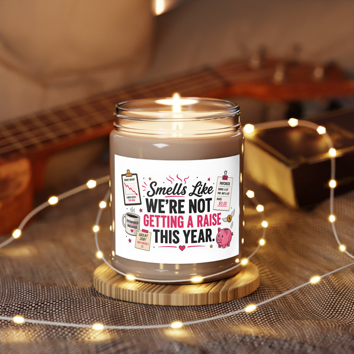 A lit candle in a clear glass jar sits on a small wooden coaster, surrounded by warm fairy lights. The candle has a white label with a humorous quote that reads 'Smells Like WE'RE NOT GETTING A RAISE THIS YEAR.